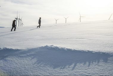 Two People Walking In The Snow With Wind Turbines