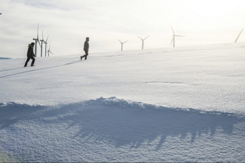 Two People Walking In The Snow With Wind Turbines